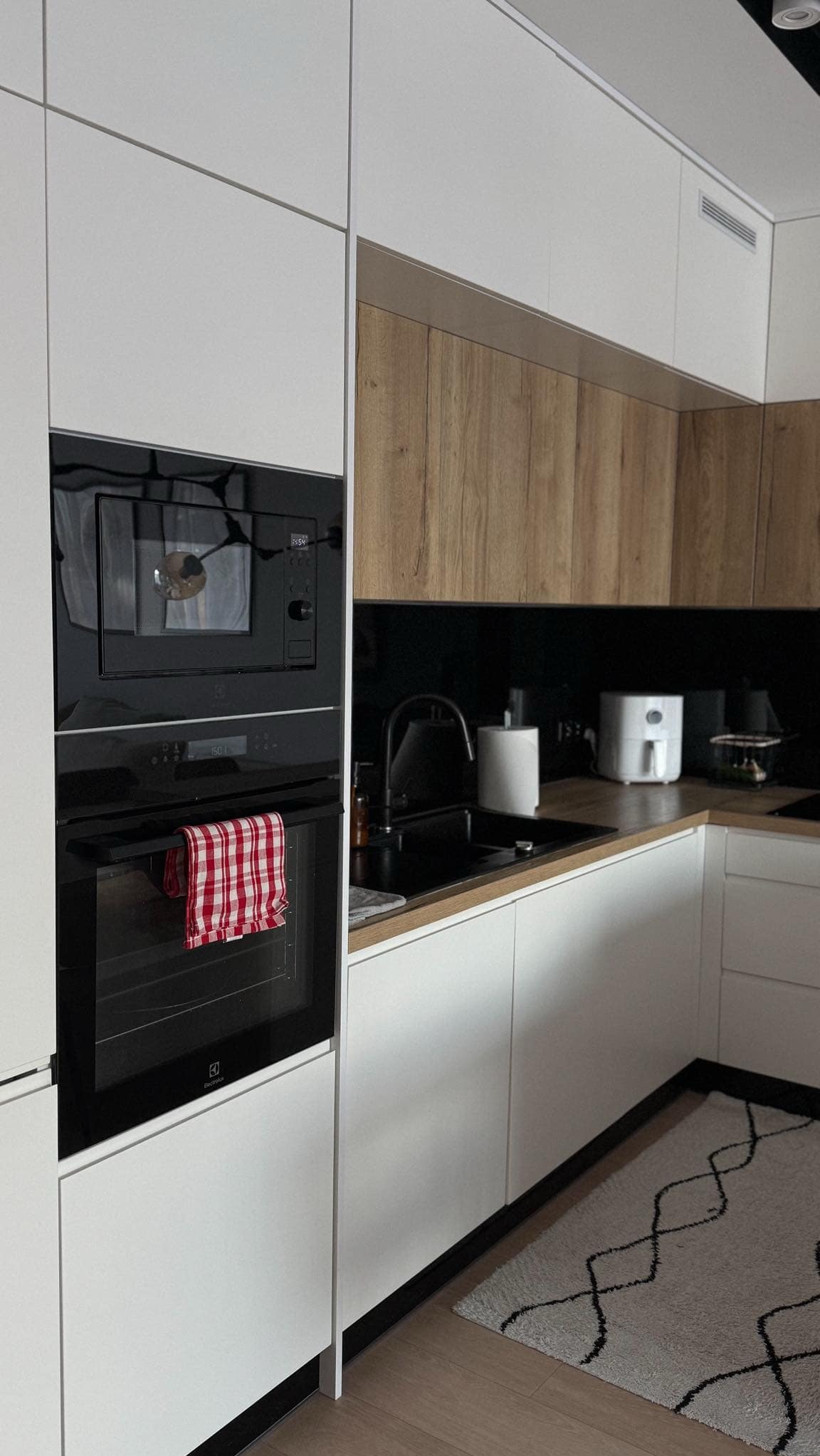 Nowoczesna kuchnia z białymi frontami, drewnianymi akcentami, czarnym blatem i piekarnikiem Electrolux z czerwoną ściereczką w kratę.