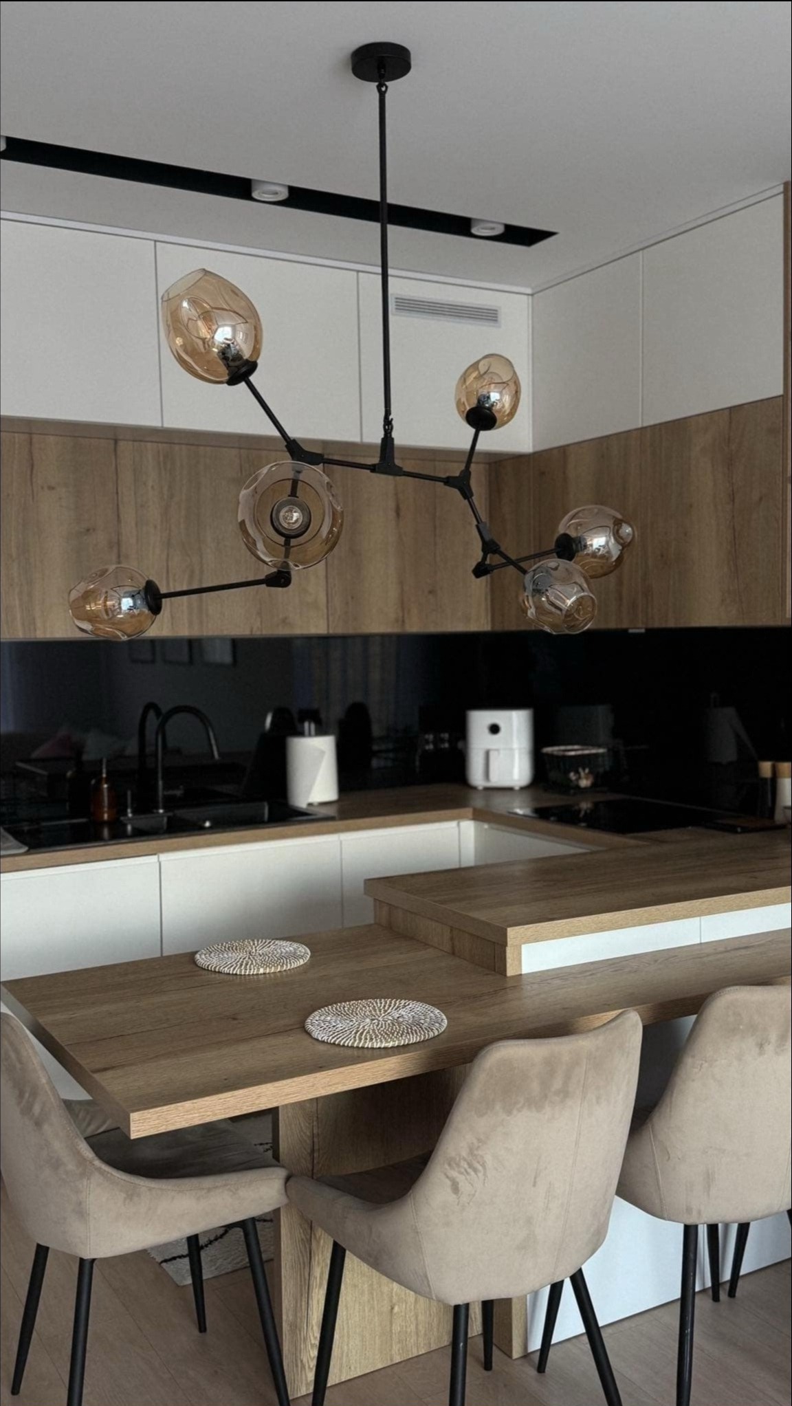 Nowoczesna kuchnia z drewnianymi i białymi szafkami, czarnym panelem nad blatem oraz designerską lampą wiszącą z bursztynowymi kloszami nad drewnianym stołem z beżowymi krzesłami.