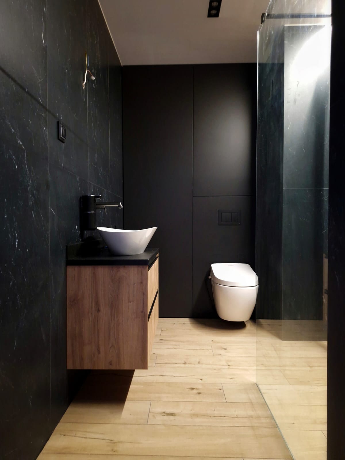Nowoczesna łazienka z czarnymi marmurowymi płytkami, drewnianą szafką pod umywalką z blatem z czarnego kamienia, białą miską umywalkową i toaletą oraz podłogą z płytek imitujących jasne drewno.