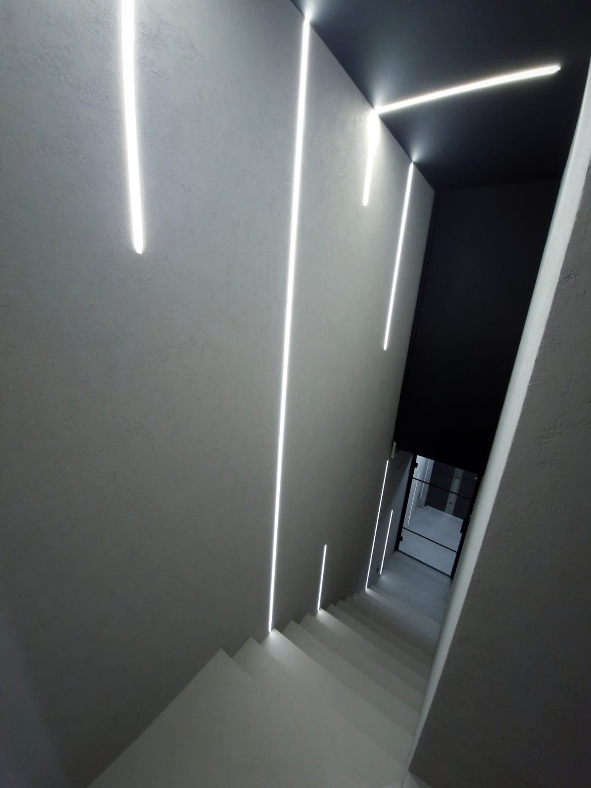 Minimalistyczne schody z oświetleniem LED wzdłuż ścian, prowadzące do pomieszczenia z przeszklonymi drzwiami, całość w odcieniach szarości i bieli.