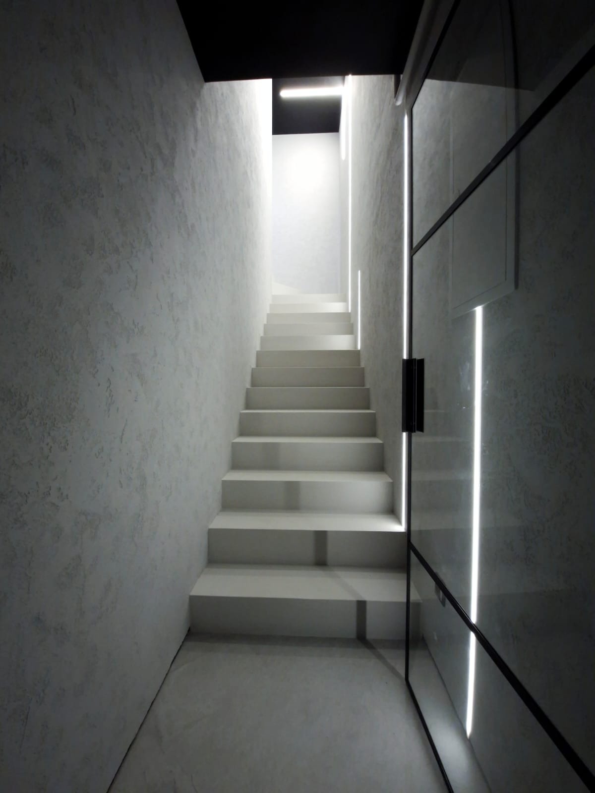 Nowoczesne schody z podświetleniem LED w Sokołowie Małopolskim, prowadzące w górę z minimalistycznym wykończeniem ścian i szklaną ścianą z czarnymi ramami.