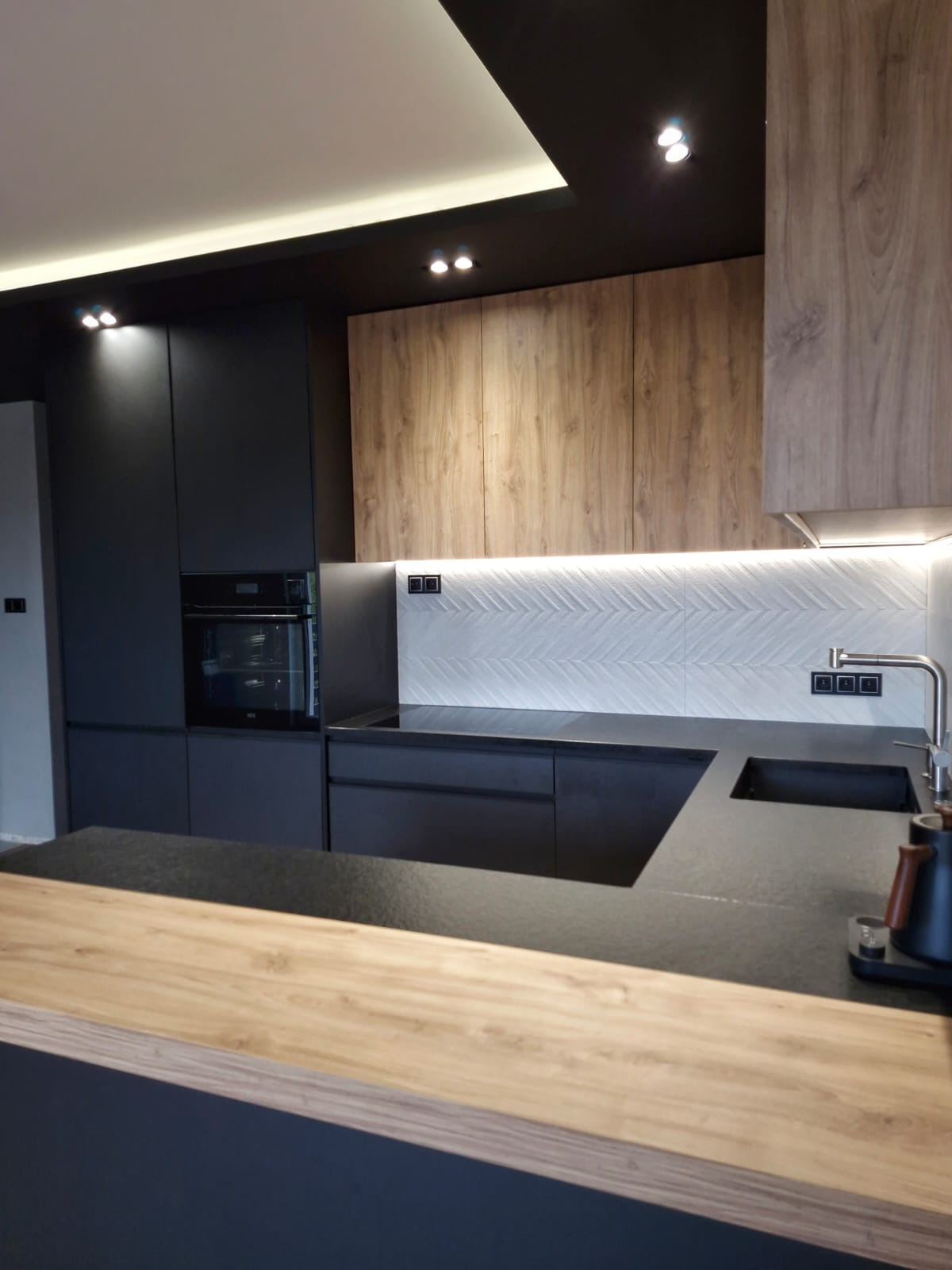 Nowoczesna kuchnia z czarnymi matowymi frontami i drewnianymi akcentami, podświetlana mozaika na ścianie, czarny blat i drewniany barek.