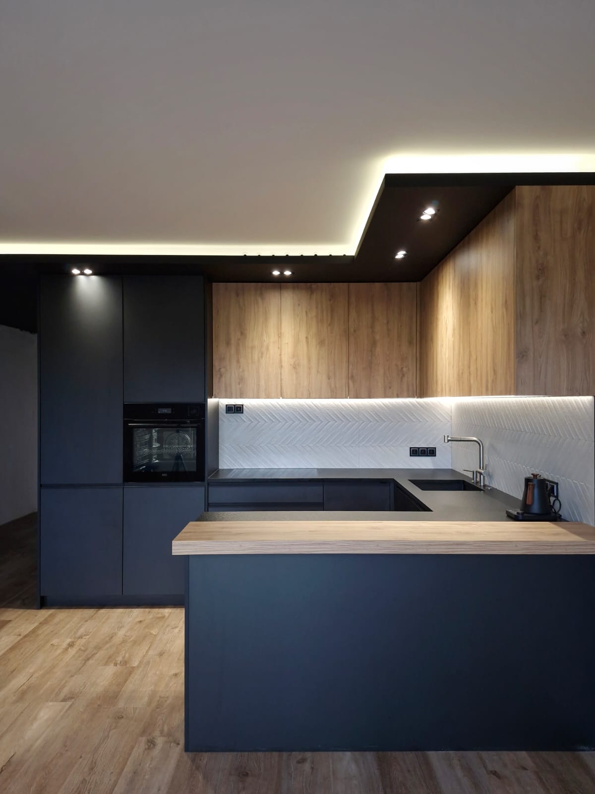 Nowoczesna kuchnia z ciemnymi szafkami i drewnianymi akcentami, oświetlenie LED podsufitowe, białe płytki w jodełkę nad blatem, drewniana podłoga.