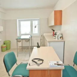 Jasne wnętrze gabinetu z beżowymi płytkami, wyposażone w stół zabiegowy ze stalową konstrukcją, biurko z turkusowymi krzesłami, na którym leży stetoskop i książka, oraz umywalkę z dozownikiem mydła.