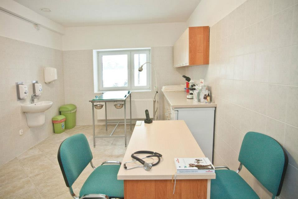 Jasne wnętrze gabinetu z beżowymi płytkami, wyposażone w stół zabiegowy ze stalową konstrukcją, biurko z turkusowymi krzesłami, na którym leży stetoskop i książka, oraz umywalkę z dozownikiem mydła.