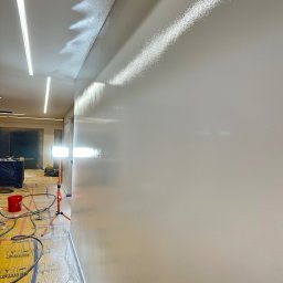 DomArt Dominik Gomółka - Gładka, świeżo pomalowana ściana w jasnym odcieniu, z widocznymi śladami światła odbijającego się od powierzchni. Na podłodze folia ochronna i narzędzia malarskie.