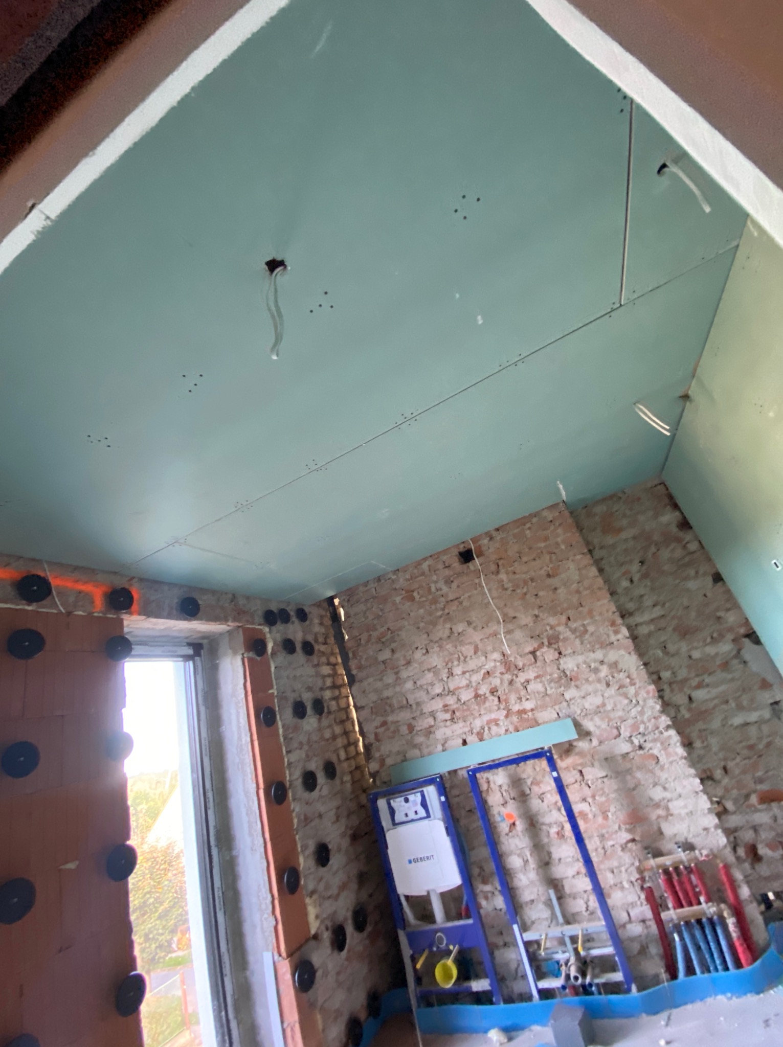 Niedokończone wnętrze z surową cegłą, ociepleniem styropianowym na ścianach, zamontowaną konstrukcją podtynkową WC marki Geberit i częściowo położonymi płytami karton-gips na suficie w kolorze...