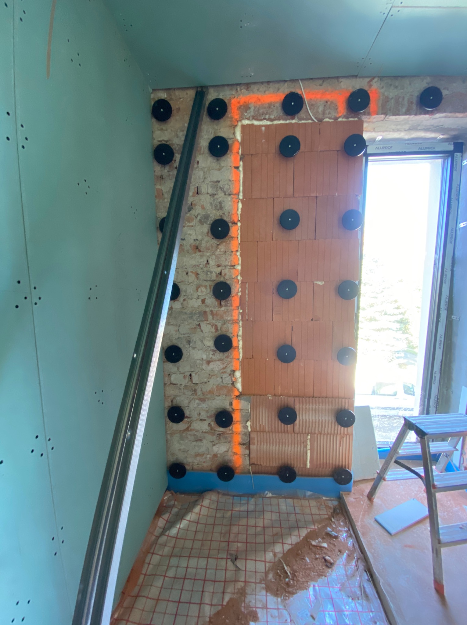 Montaż ściany z cegły z użyciem łączników talerzykowych, widoczna konstrukcja metalowa, przygotowana pod płyty kartonowo-gipsowe, na podłodze instalacja ogrzewania podłogowego.