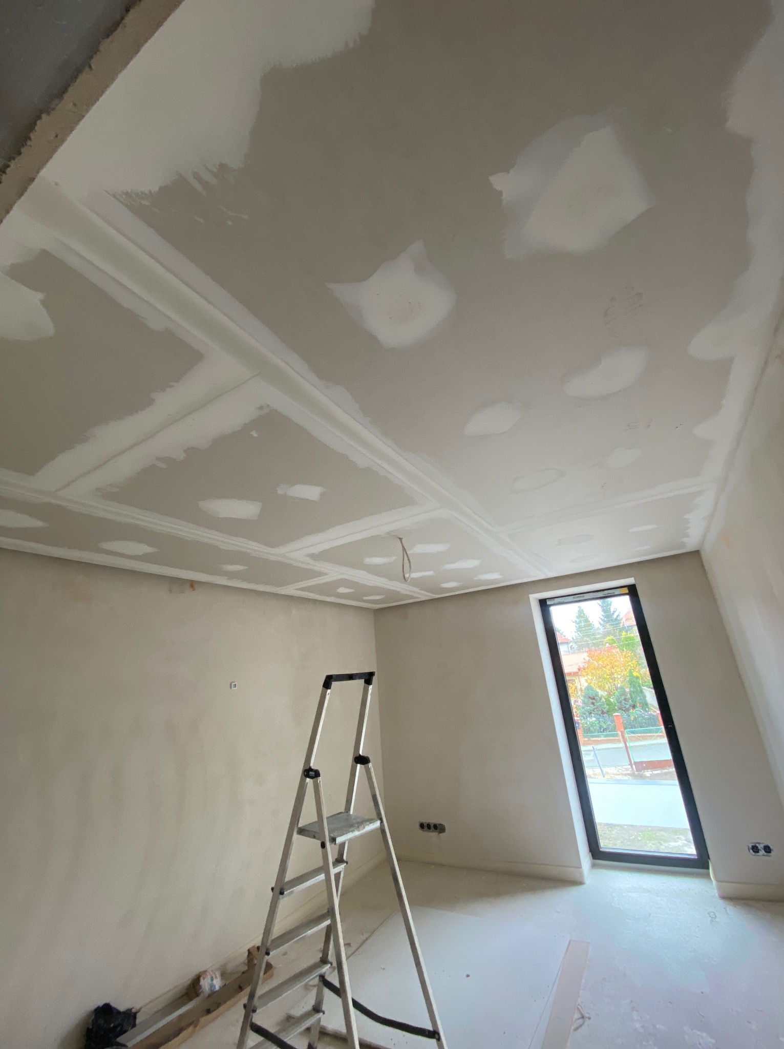 Pokój w trakcie prac wykończeniowych: sufit z płyt kartonowo-gipsowych z dekoracyjnymi listwami i widocznymi spoinami, ściany po gładzi, aluminiowa drabina rozstawiona na podłodze, widok...