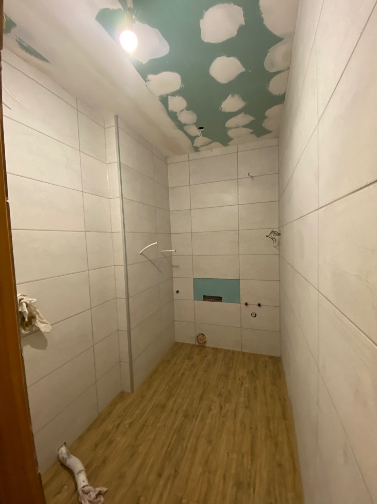 Wnętrze łazienki w trakcie prac: białe płytki na ścianach, drewnopodobna podłoga, sufit z widocznymi łatami gipsu i zieloną płytą, przygotowane punkty instalacji wodnej.