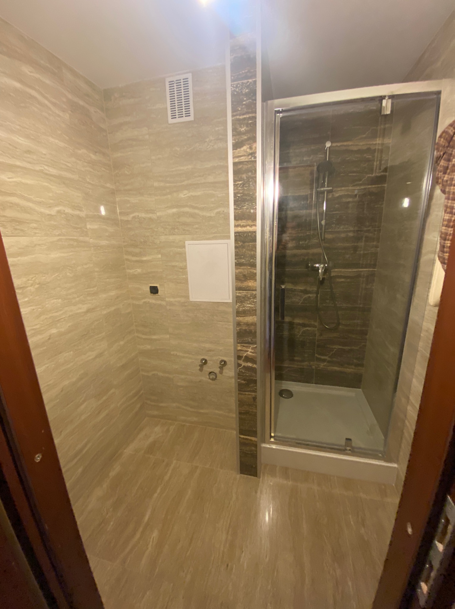 Wykończona łazienka z kabiną prysznicową i beżowymi płytkami imitującymi marmur, widok zza uchylonych drzwi.