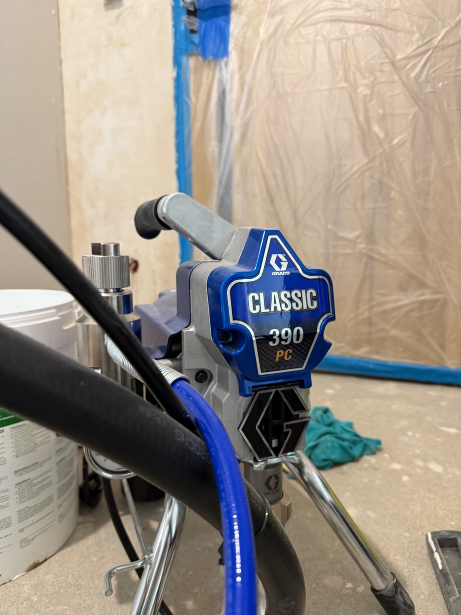 Profesjonalny agregat malarski Graco Classic 390 PC gotowy do pracy, zabezpieczone folią ściany w tle, widoczny wiadro z farbą. Sprzęt malarski w zbliżeniu.