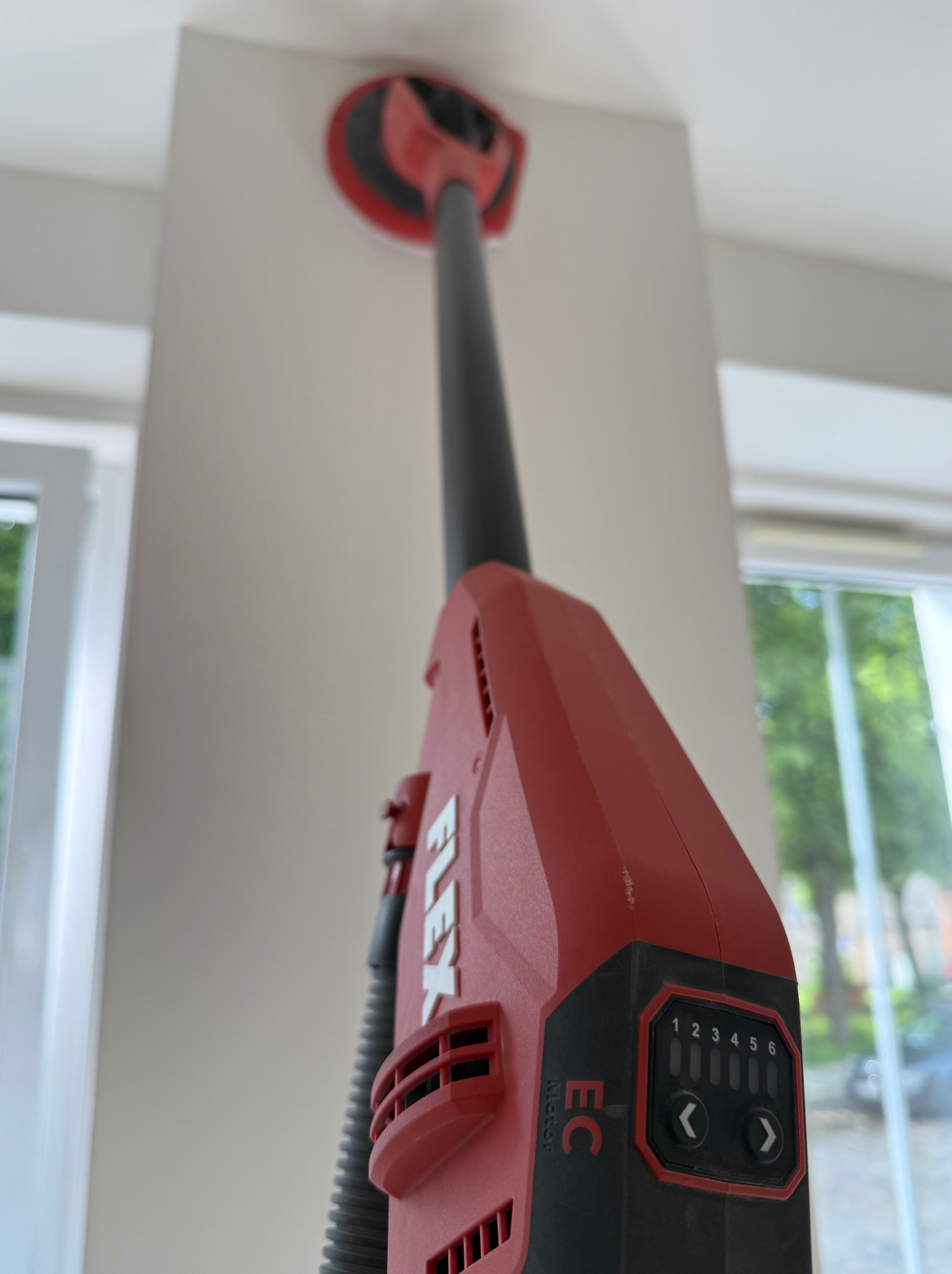 Szlifierka teleskopowa Flex do gładzi, widok z dołu na obrabianą ścianę i okno w tle. Urządzenie z regulacją obrotów, gotowe do pracy.