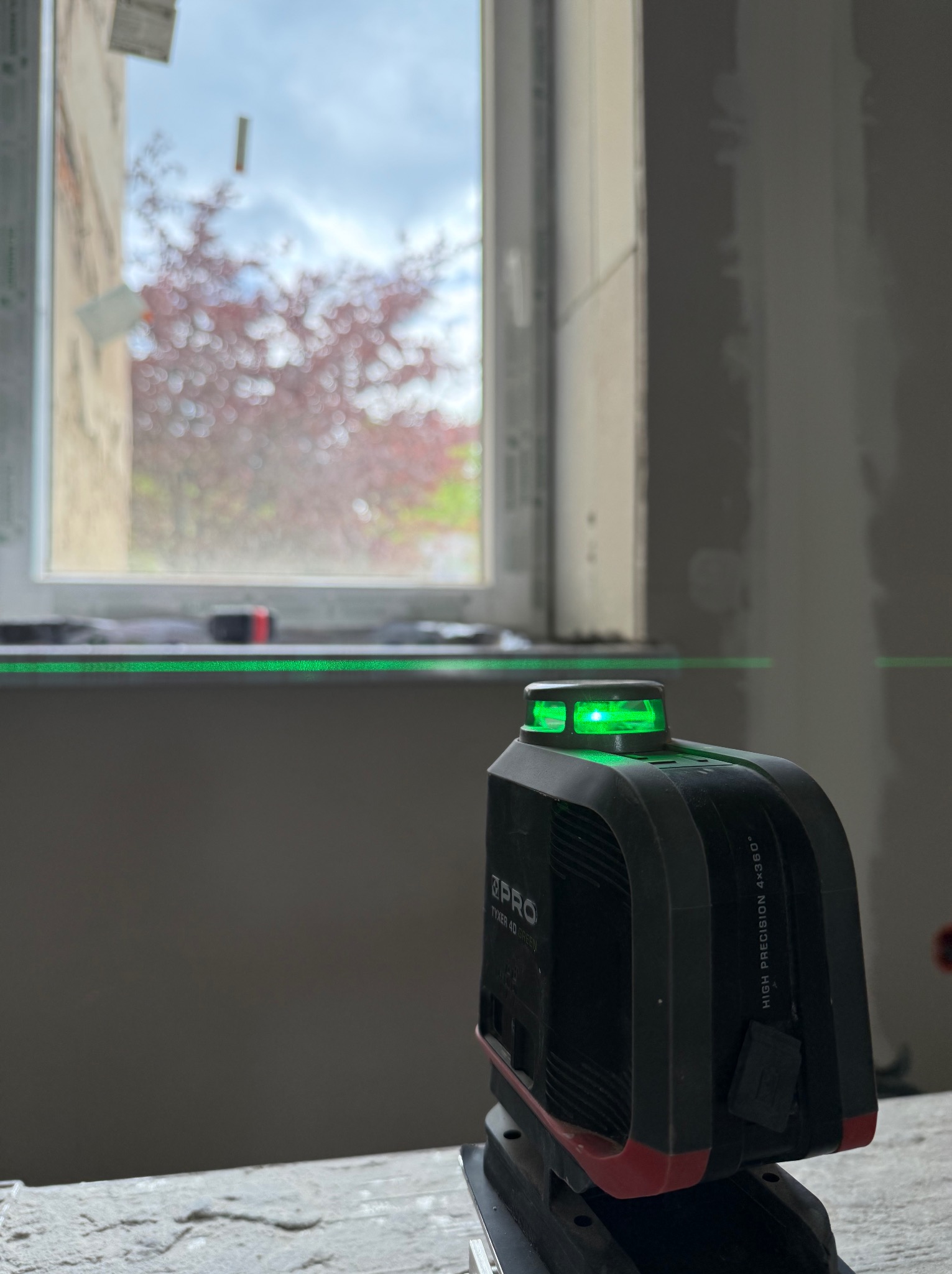 Laser krzyżowy emituje zielone linie na ścianie i oknie podczas prac wykończeniowych. Widoczny fragment ściany z nałożoną gładzią. W tle drzewo za oknem.