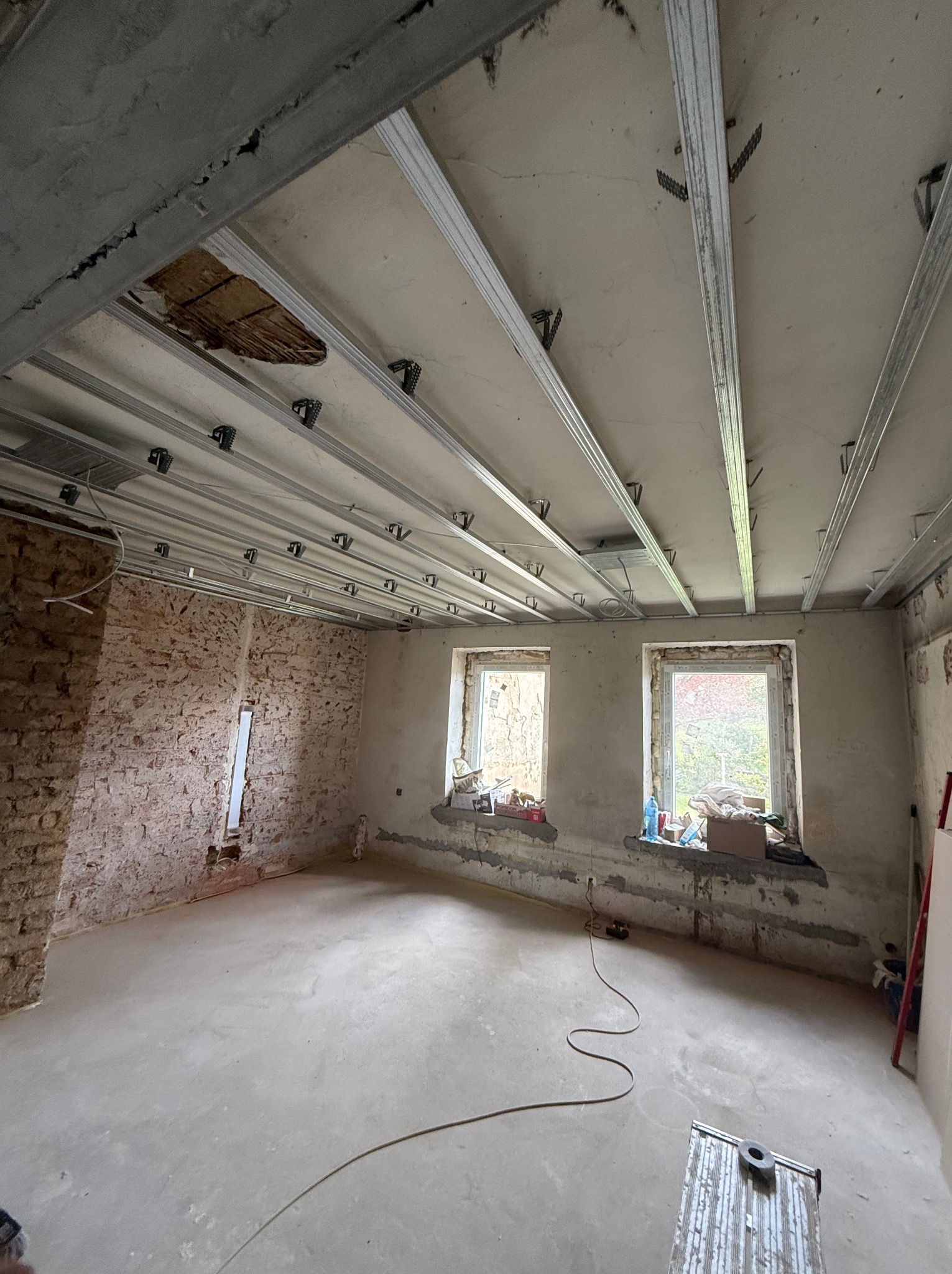 Montaż stelaża sufitu podwieszanego z płyt g-k w Kamiennej Górze. Widoczne profile i mocowania, surowy stan ścian i podłogi, dwa okna. Prace remontowe w toku.