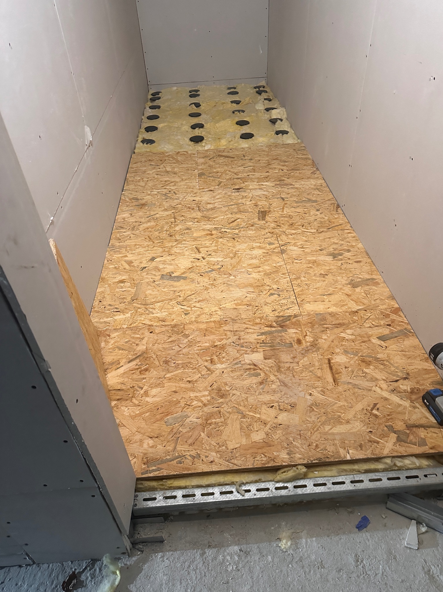 Montaż podłogi z płyt OSB na legarach z izolacją w wąskim pomieszczeniu. Ściany wykończone płytami gipsowo-kartonowymi. Widoczne elementy konstrukcyjne.