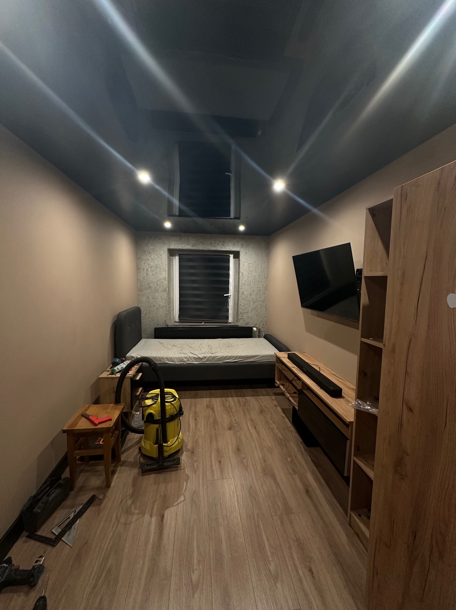 Pokój po remoncie: ciemny sufit napinany z oświetleniem punktowym, parkiet, meble. Na podłodze narzędzia i odkurzacz. Widok na ścianę z oknem i zamontowany telewizor.
