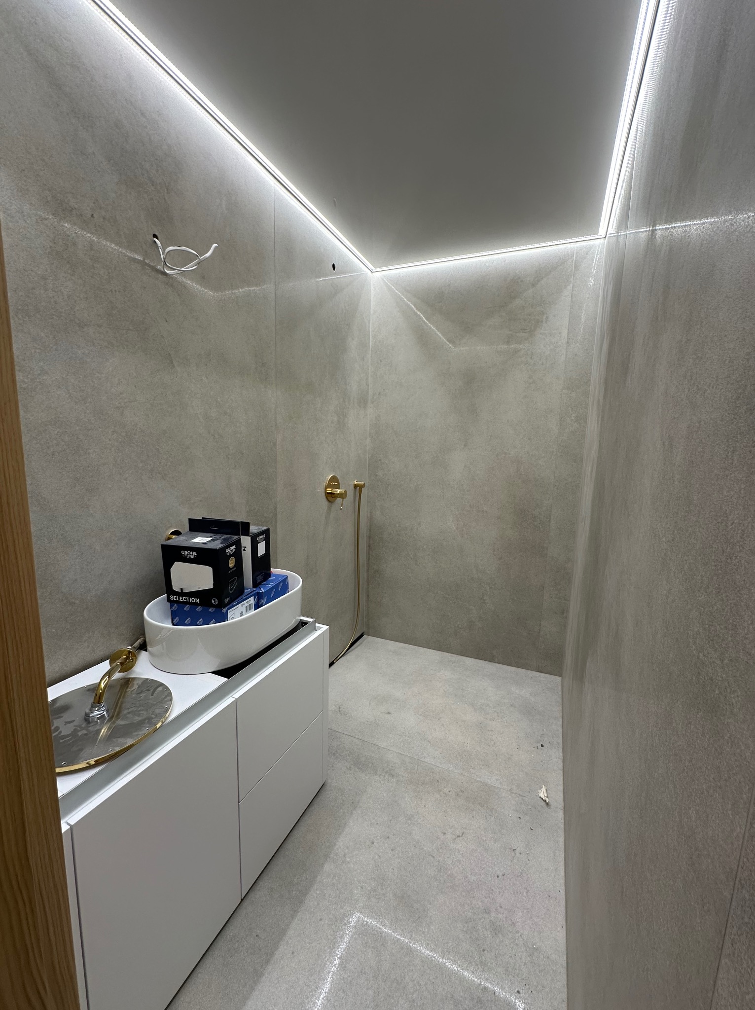 Nowoczesna łazienka w szarościach z oświetleniem LED, białą szafką pod umywalkę i złotą armaturą. Widoczne pudełka z armaturą GROHE.