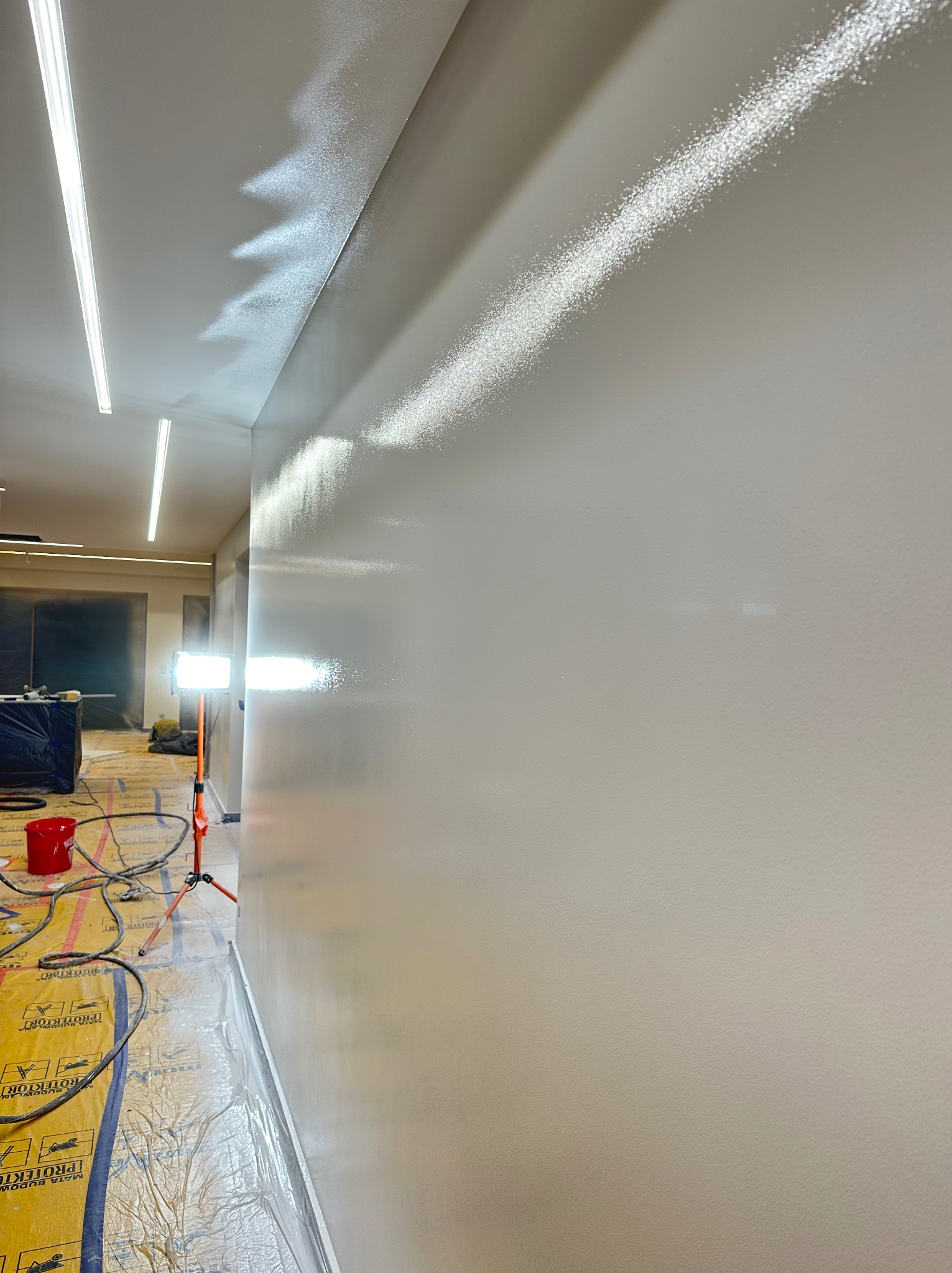 Gładka, świeżo pomalowana ściana w jasnym odcieniu, z widocznymi śladami światła odbijającego się od powierzchni. Na podłodze folia ochronna i narzędzia malarskie.