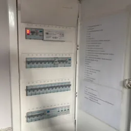 Otwarta skrzynka elektryczna z widocznymi bezpiecznikami i listwą zasilającą, obok naklejona kartka z opisem obwodów.