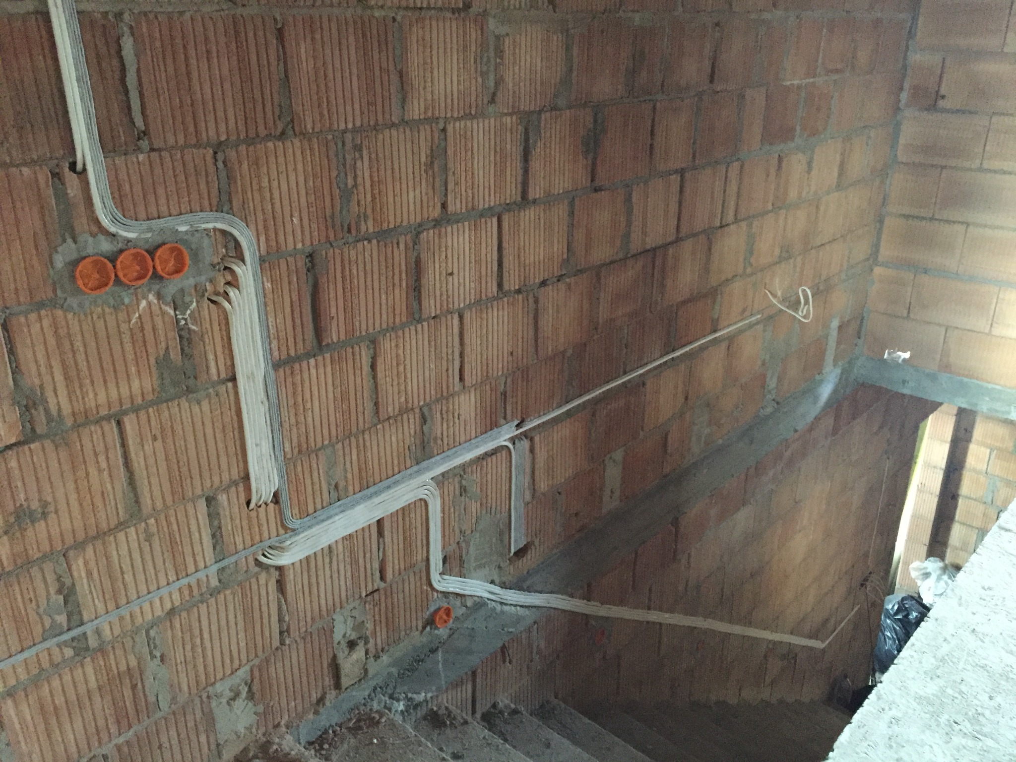 Instalacja elektryczna w surowym wnętrzu: kable prowadzone po ścianie z cegły dziurawki, pomarańczowe puszki pod gniazdka, widok na schody i otwór w stropie.