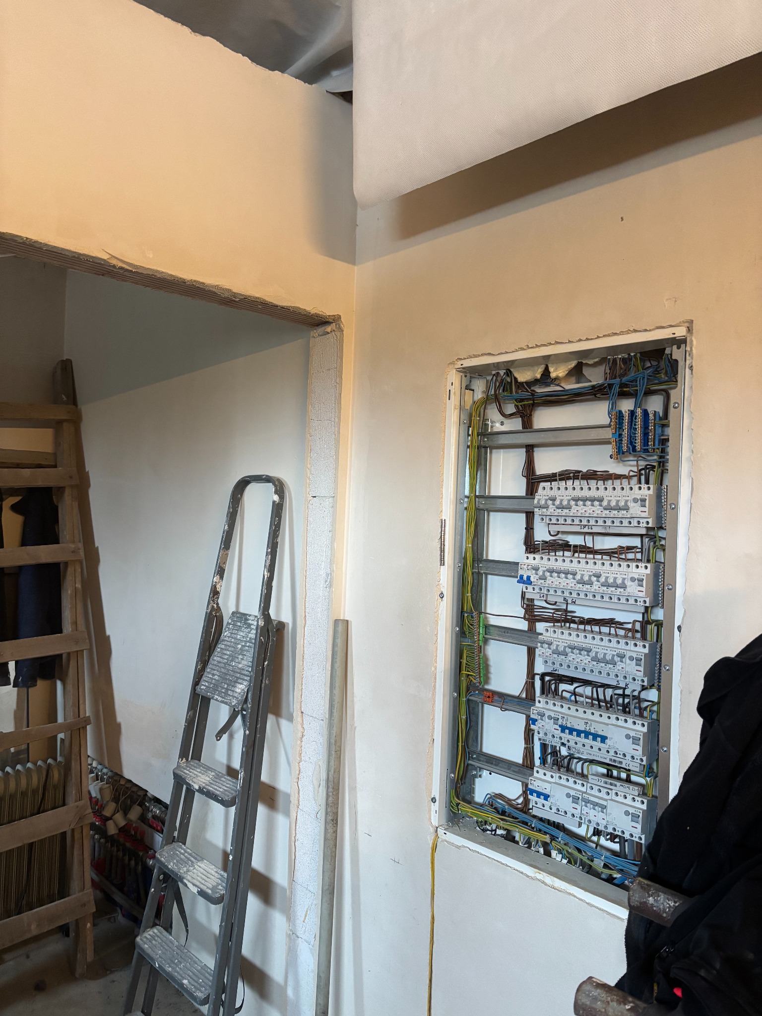 Rozdzielnica elektryczna w trakcie montażu w surowym wnętrzu, widoczne przewody, zabezpieczenia, drabiny i ściany w trakcie wykończenia.