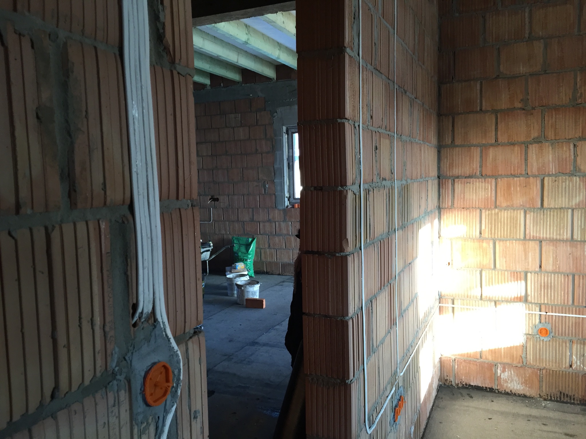 Wewnętrzne ściany budynku w trakcie budowy, z widocznymi przewodami elektrycznymi w białych listwach i pomarańczowymi puszkami instalacyjnymi, murowane z czerwonej cegły dziurawki.