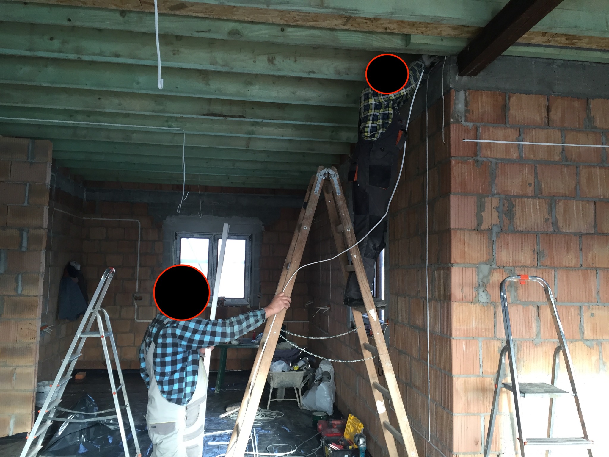 Dwóch elektryków na drabinach montuje przewody elektryczne w surowym wnętrzu budynku z widocznymi cegłami i drewnianą konstrukcją stropu.