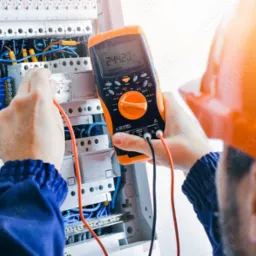 Elektryk w kasku, mierzący napięcie w skrzynce rozdzielczej za pomocą multimetru cyfrowego; widoczne przewody, bezpieczniki i wyświetlacz z wartością pomiaru.