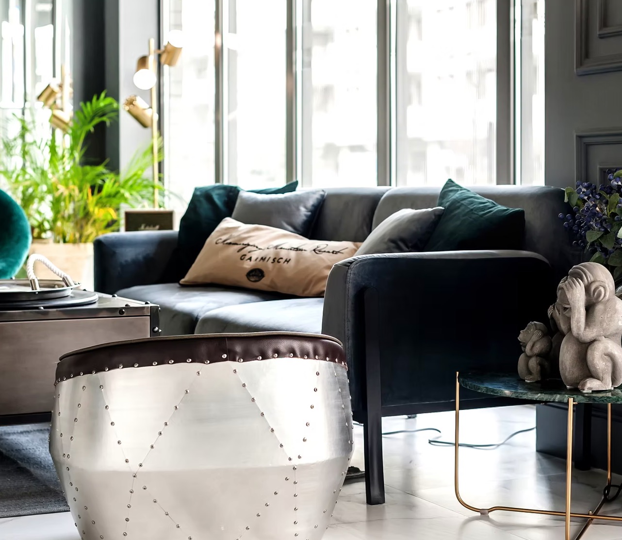 Stylowy salon z szarą sofą, zielonymi poduszkami i metalowym pufem. W tle okno i roślina doniczkowa, a na stoliku dekoracyjne figurki małp.