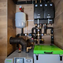 Zmontowana kotłownia z piecem na pellet, zbiornikiem wyrównawczym i osprzętem, widoczne rury instalacji grzewczej, manometry i zawory, całość w pomieszczeniu wyłożonym płytkami imitującymi drewno.