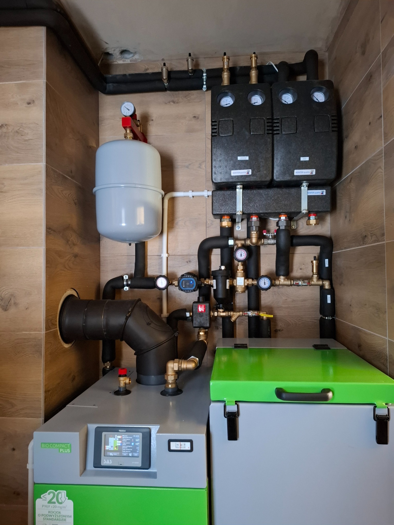 Zmontowana kotłownia z piecem na pellet, zbiornikiem wyrównawczym i osprzętem, widoczne rury instalacji grzewczej, manometry i zawory, całość w pomieszczeniu wyłożonym płytkami imitującymi drewno.