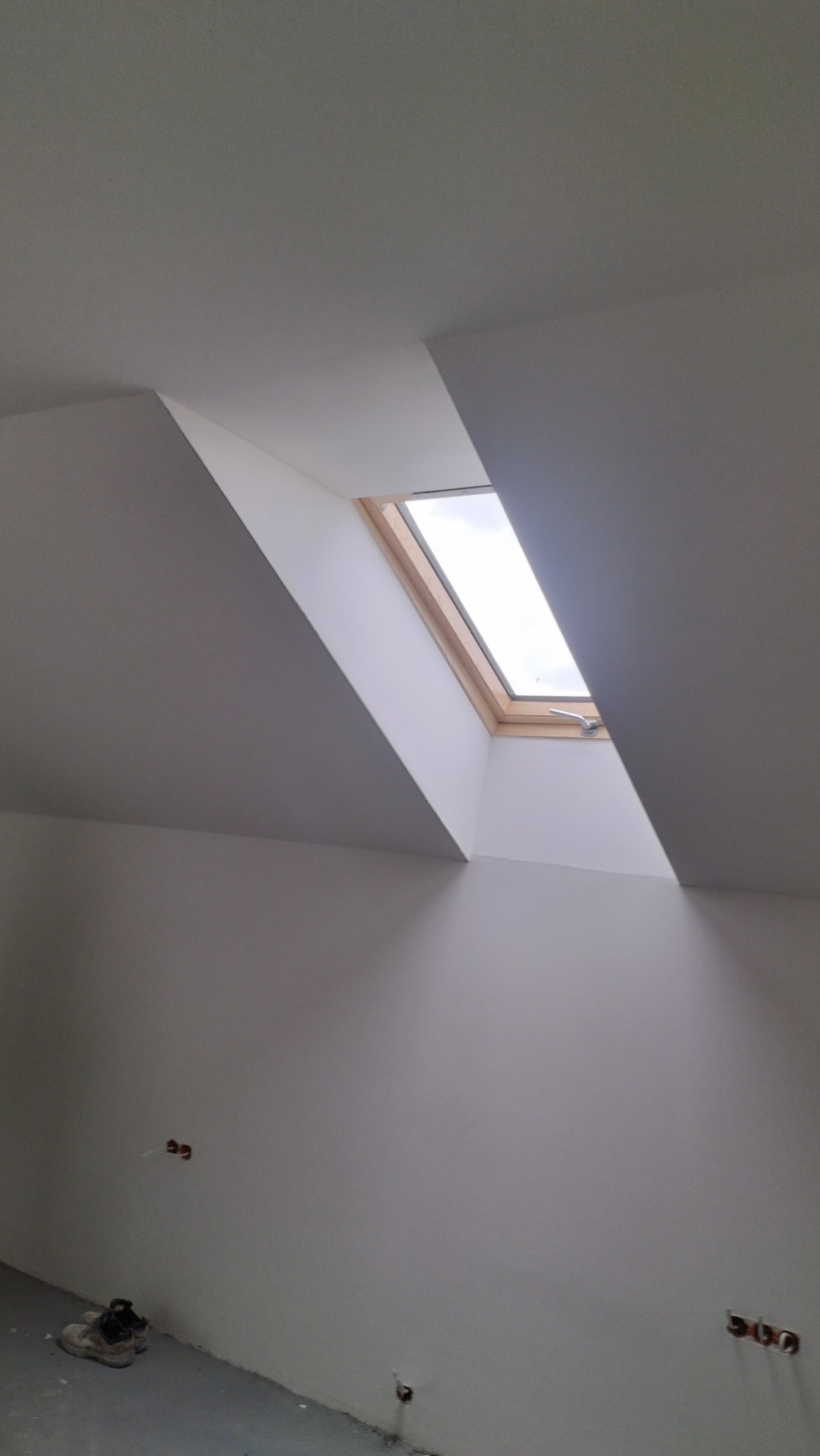 Poddasze po remoncie: białe ściany, okno dachowe z drewnianą ramą, gładzie wykończone, widoczne gniazdka elektryczne. Buty robocze w rogu.