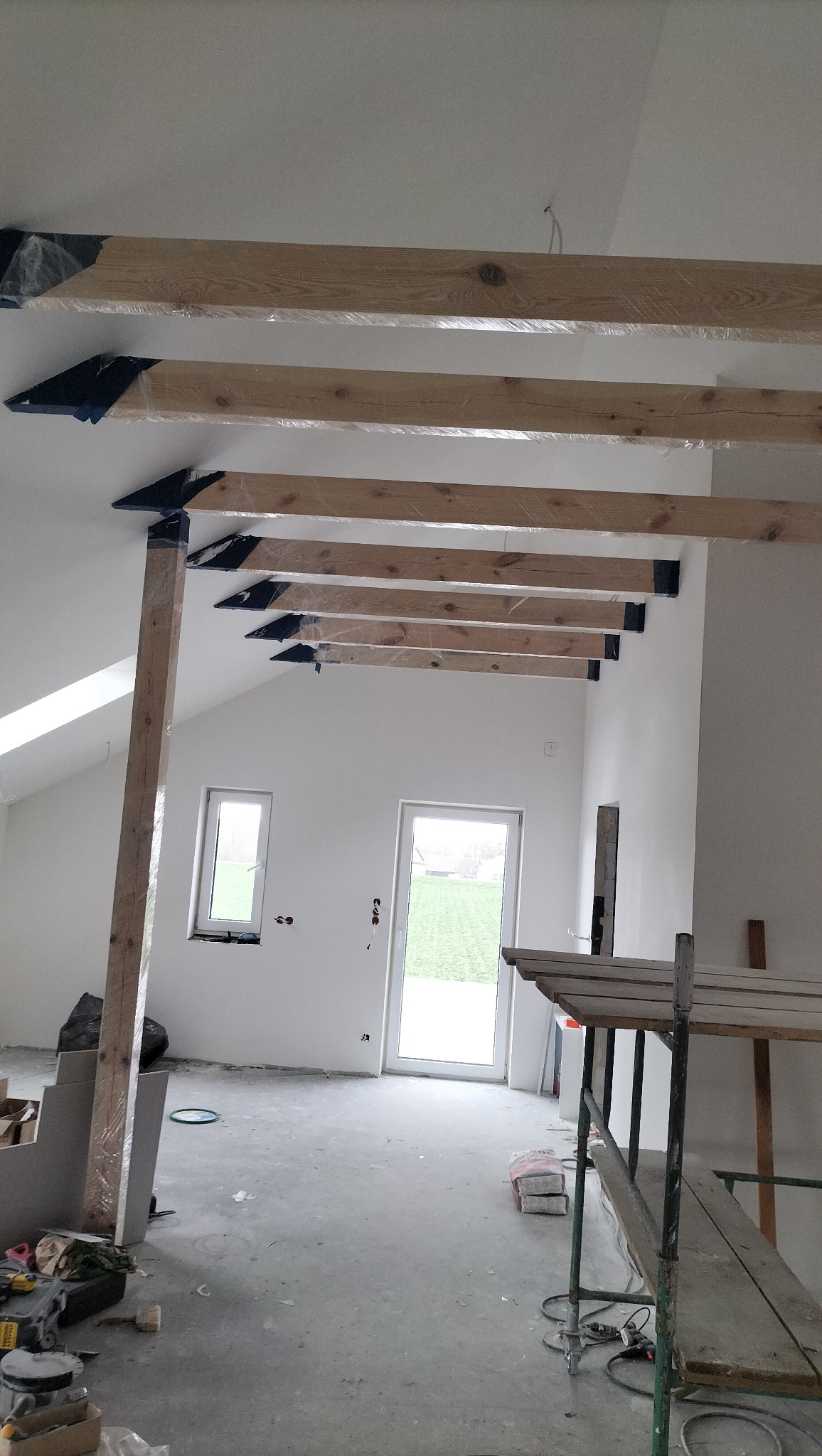 Drewniane belki stropowe, zabezpieczone folią, zamontowane w surowym wnętrzu z oknami i drzwiami. Widoczne rusztowanie i materiały budowlane na podłodze.
