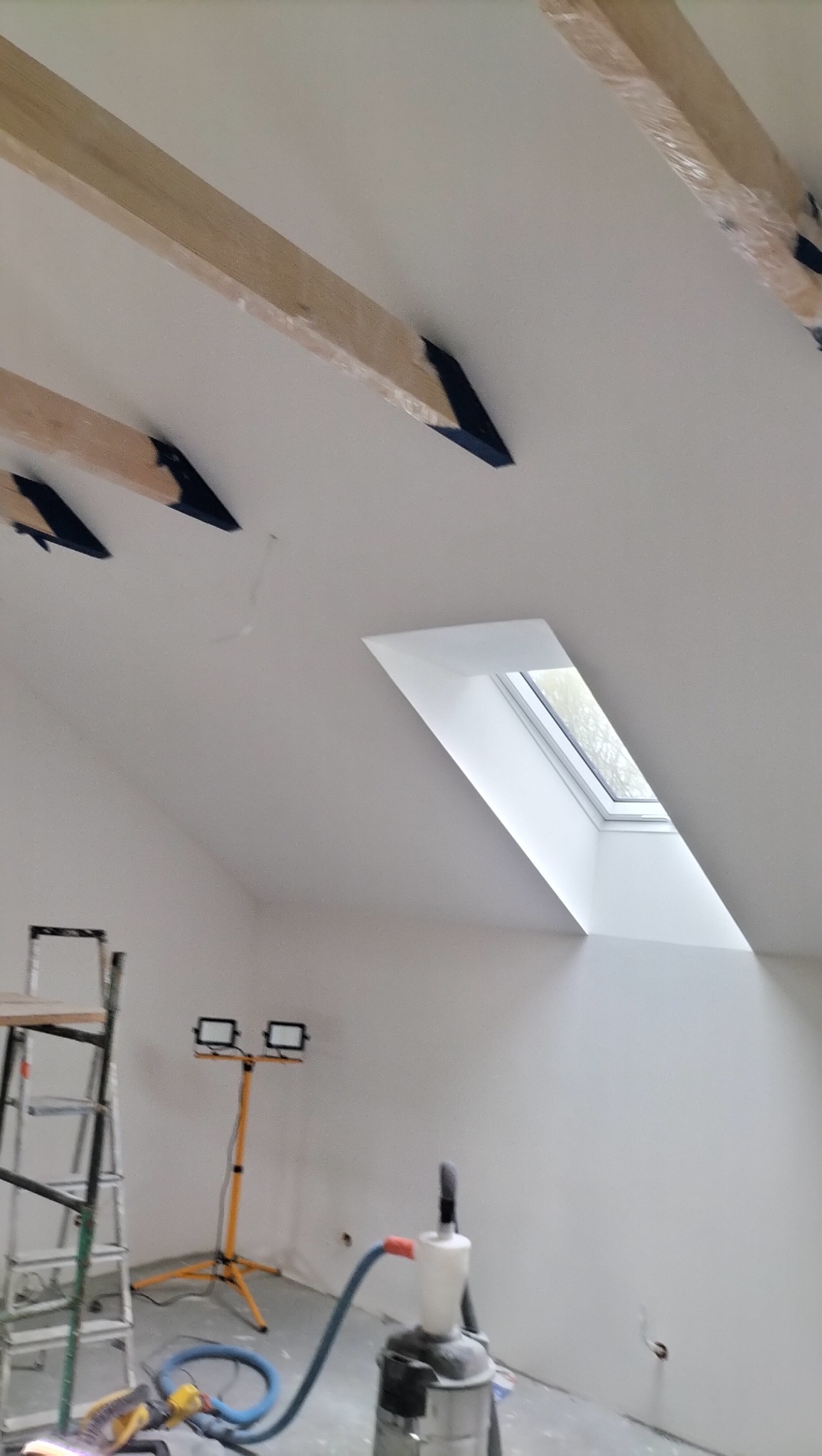 Białe poddasze ze skośnym oknem dachowym i drewnianymi belkami zabezpieczonymi folią. Widoczny sprzęt budowlany: drabina, lampa i odkurzacz przemysłowy.