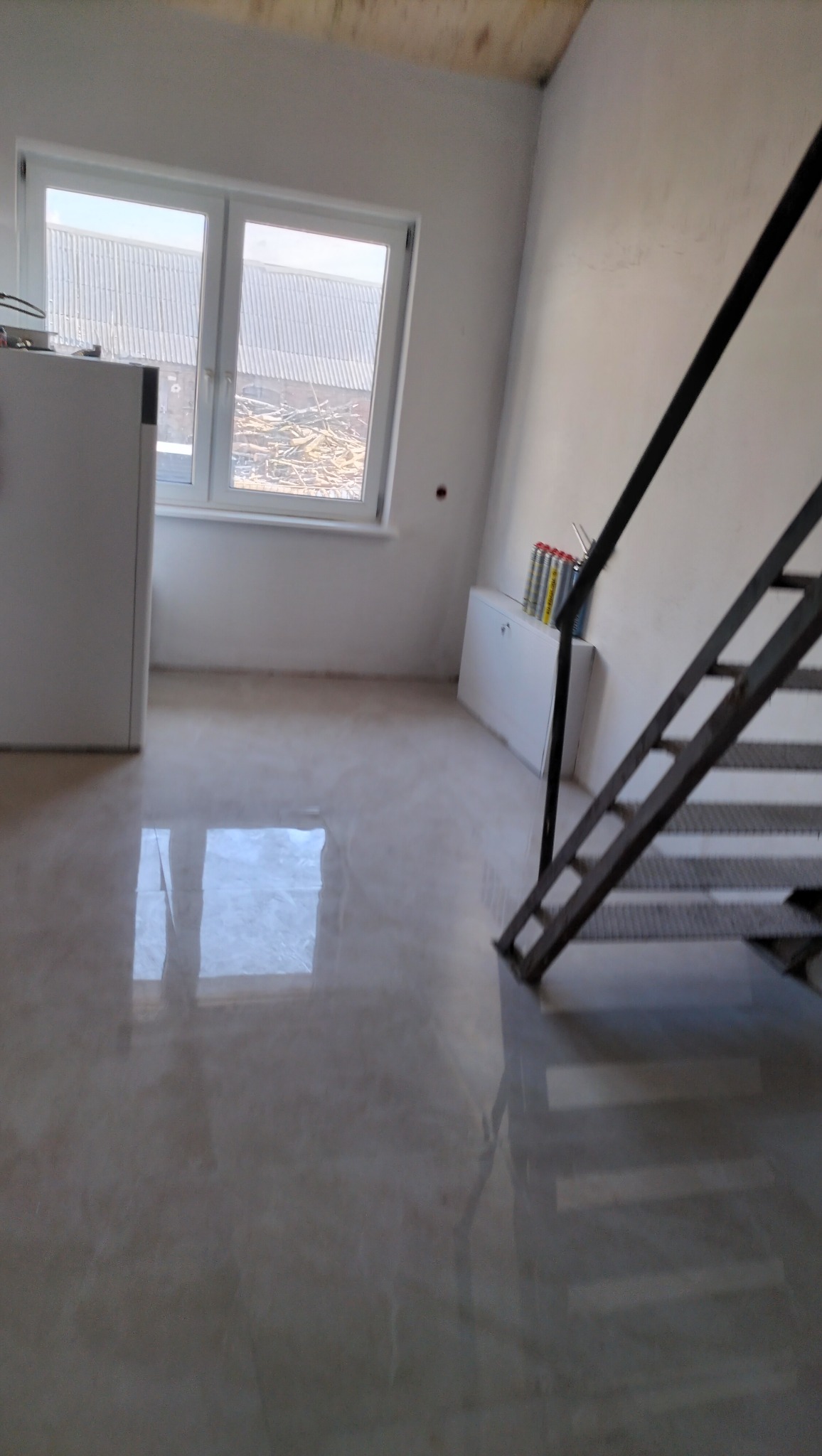 Błyszcząca, jasna posadzka w nowym wnętrzu z oknem i metalowymi schodami. Widoczne materiały budowlane przy ścianie. Minimalistyczny design, surowy stan wykończenia.