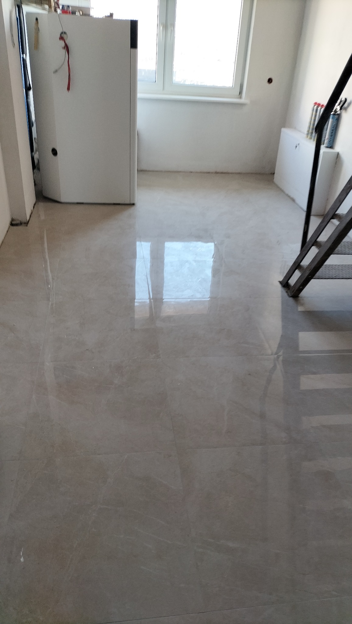 Błyszcząca, beżowa podłoga z płytek po remoncie, odbijająca światło okna. Widoczny fragment schodów i białe ściany. Minimalistyczne wykończenie wnętrza.