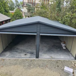 Nowo postawiony, dwustanowiskowy garaż z szarej blachy, z otwartymi wrotami, widok z zewnątrz na żwirowe podłoże i częściowo zarośnięte otoczenie.