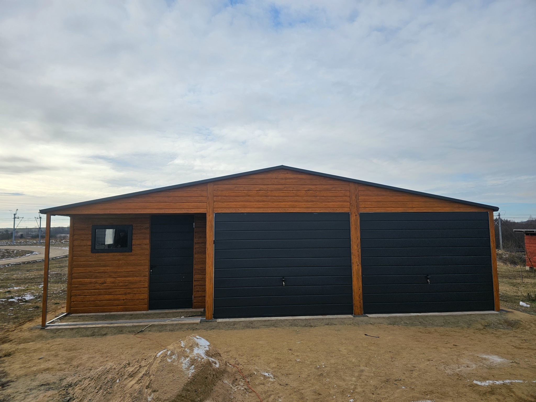 Gotowy, drewnopodobny garaż z dwiema bramami segmentowymi w kolorze grafitowym i bocznym wejściem, na nierównym terenie.