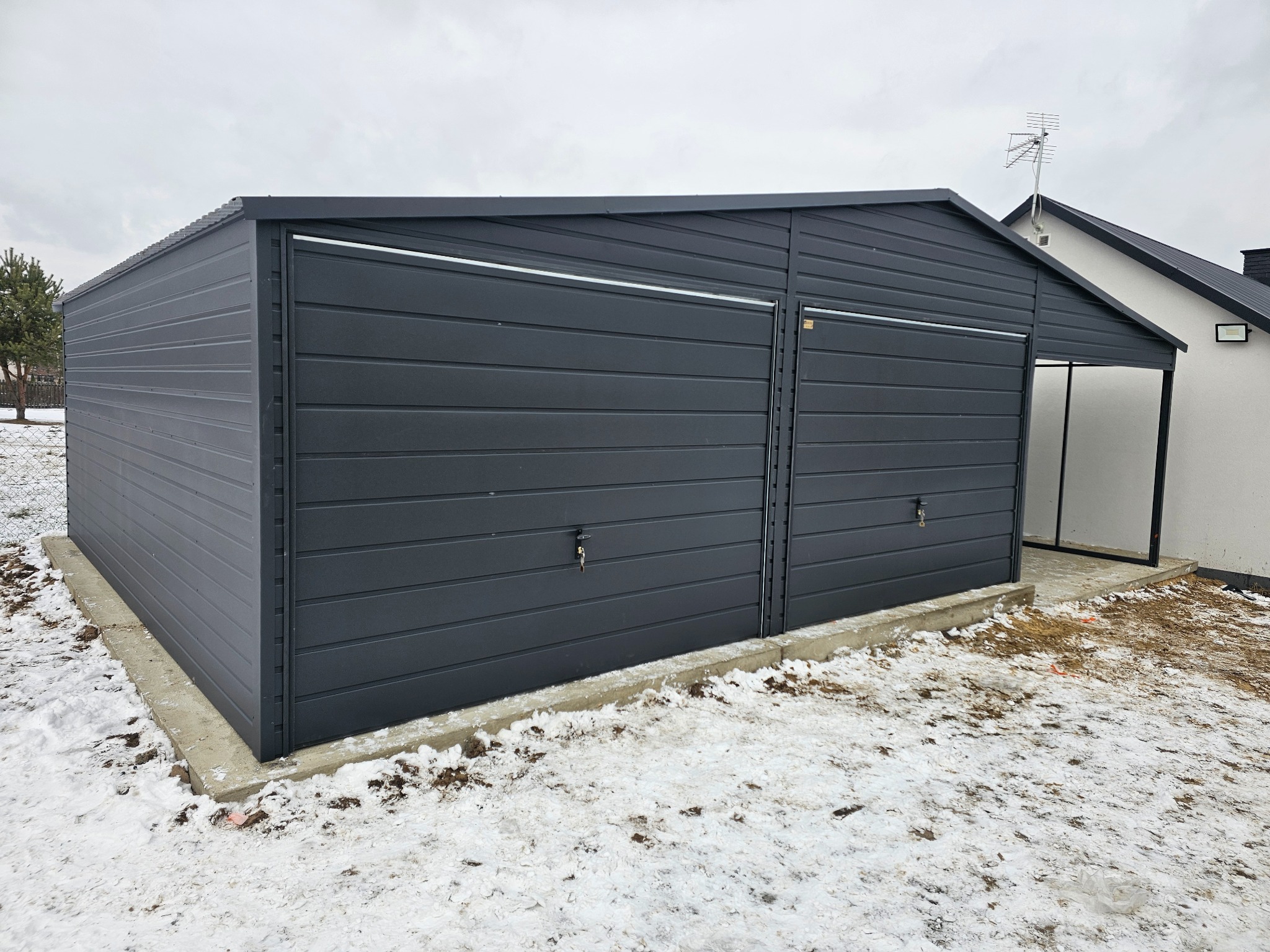 Nowoczesny, ciemnoszary garaż z blachy falistej, dwustanowiskowy, z widocznymi zamkami w drzwiach, posadowiony na betonowej wylewce, na tle zaśnieżonego terenu i domu z anteną.