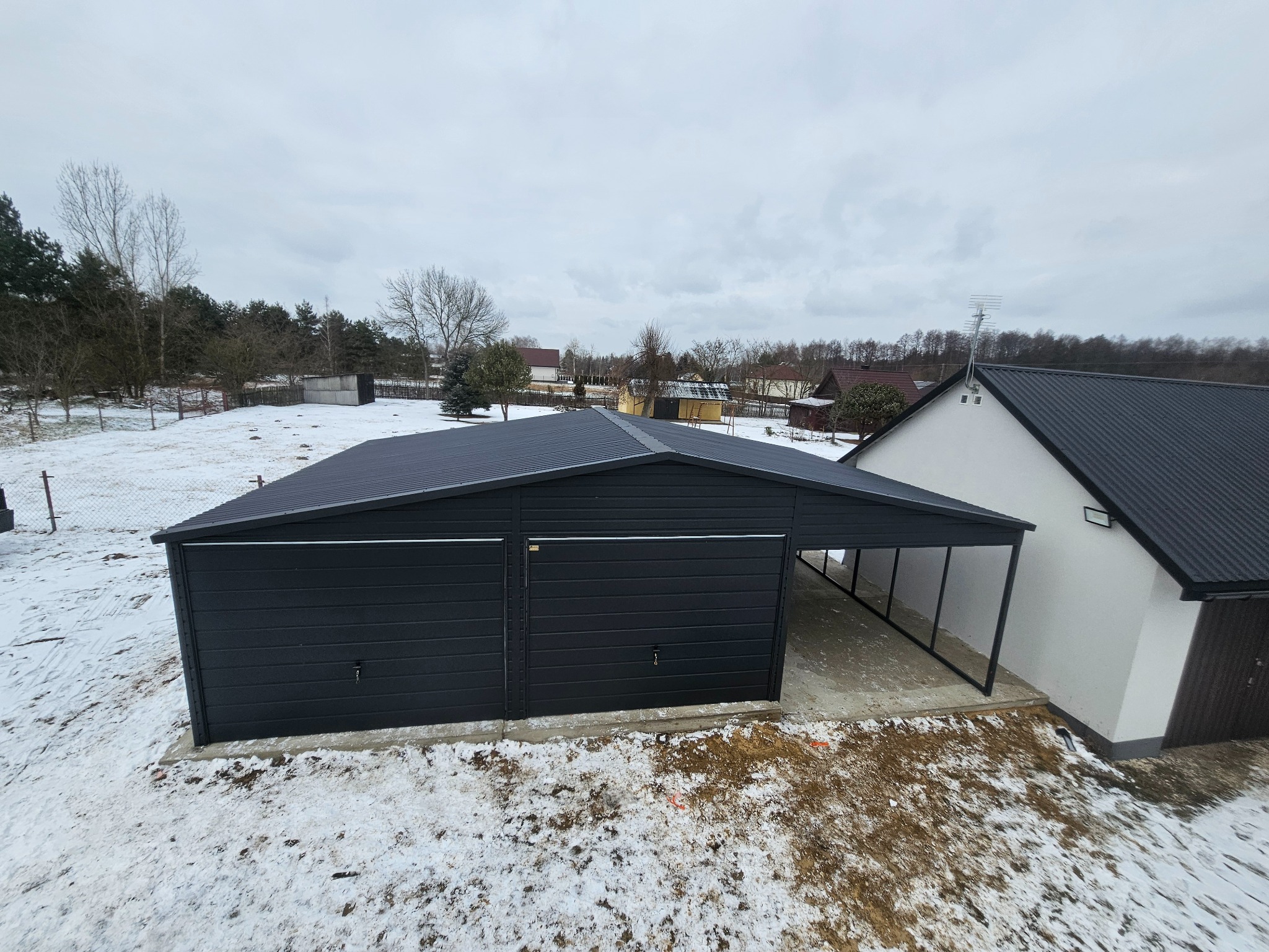 Czarny, dwustanowiskowy garaż z blachy trapezowej z wiatą, na betonowej wylewce, w zimowym krajobrazie z widokiem na dom i drzewa.