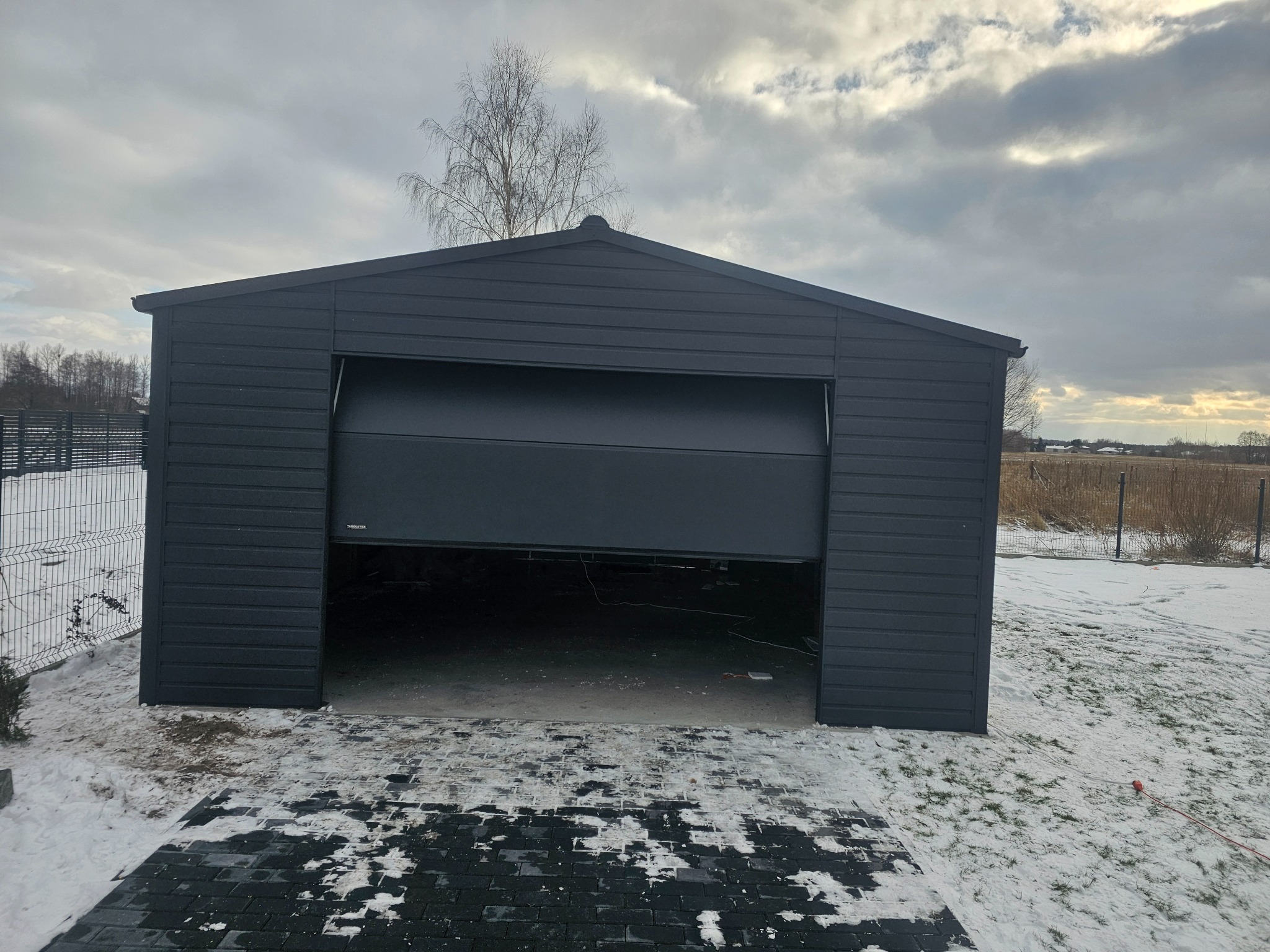 Grafitowy garaż blaszany z częściowo otwartą bramą segmentową, na posesji z kostką brukową i resztkami śniegu, widok na pole i pochmurne niebo.