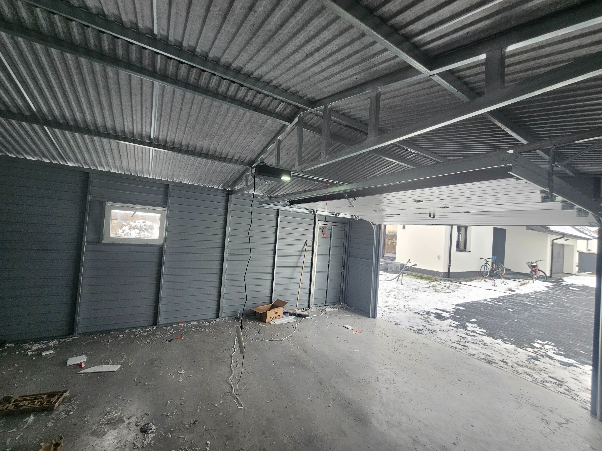 Wnętrze szarego garażu blaszanego z metalową konstrukcją dachu, widoczne narzędzia i nieporządek na podłodze, brama garażowa otwarta na ośnieżony podjazd.
