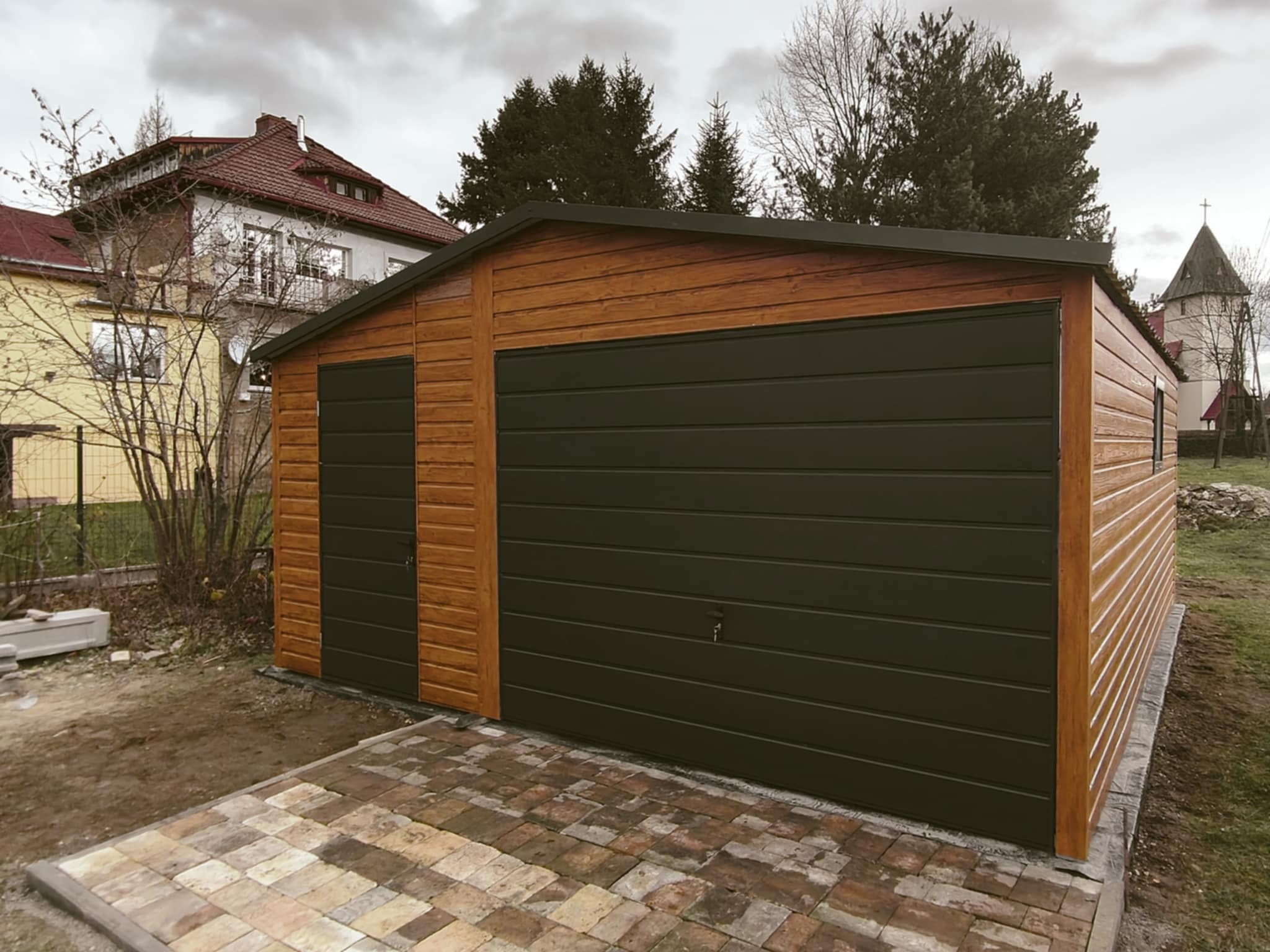 Gotowy garaż z brązowego drewna z dwiema bramami w kolorze grafitowym, ułożony na kostce brukowej, w tle dom i kościół.