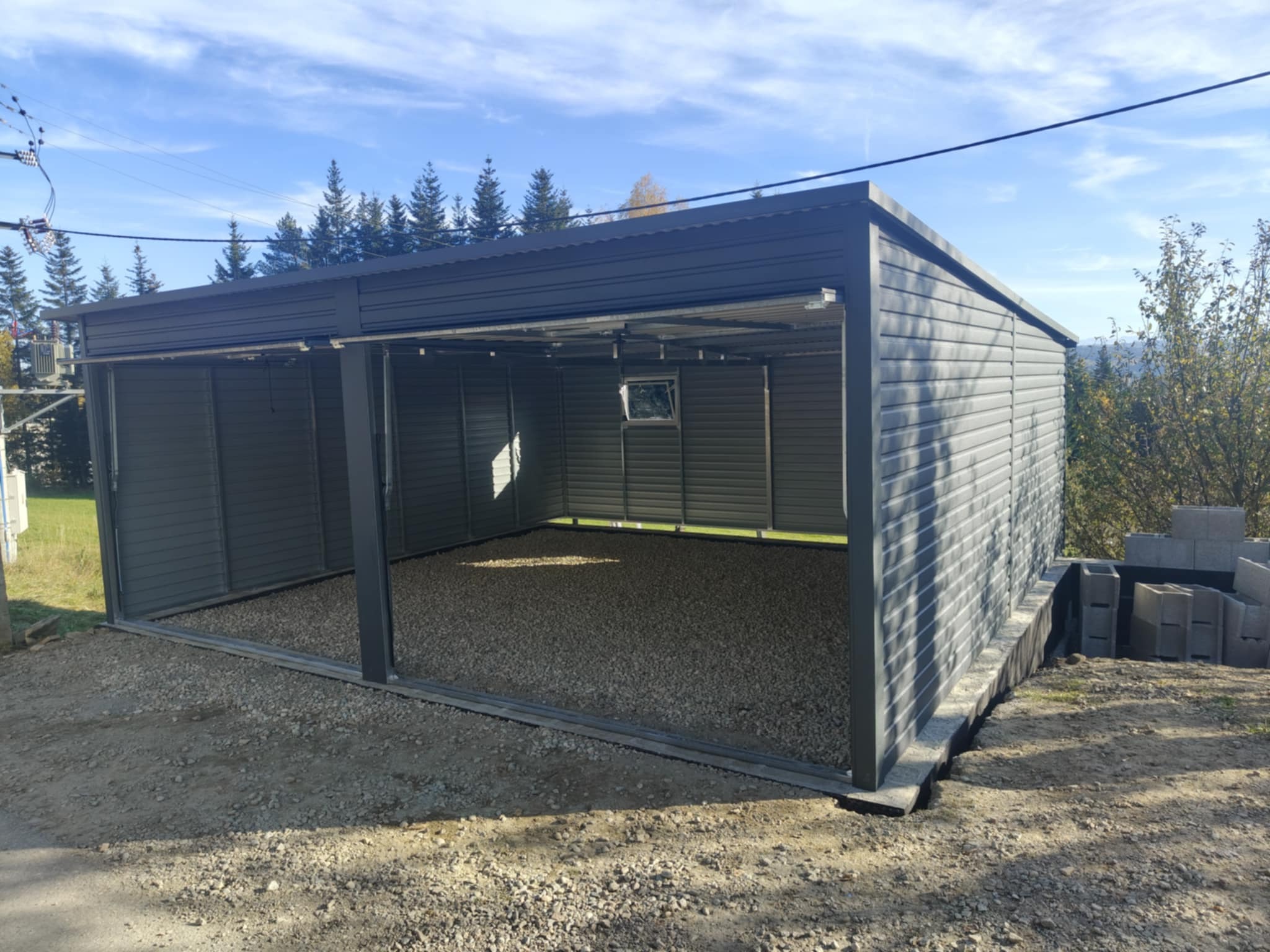 Szary, dwustanowiskowy garaż z blachy falistej na podłożu z kamyków, widok zewnętrzny w słoneczny dzień, z widocznymi drzewami w tle i stertą bloczków betonowych z boku.