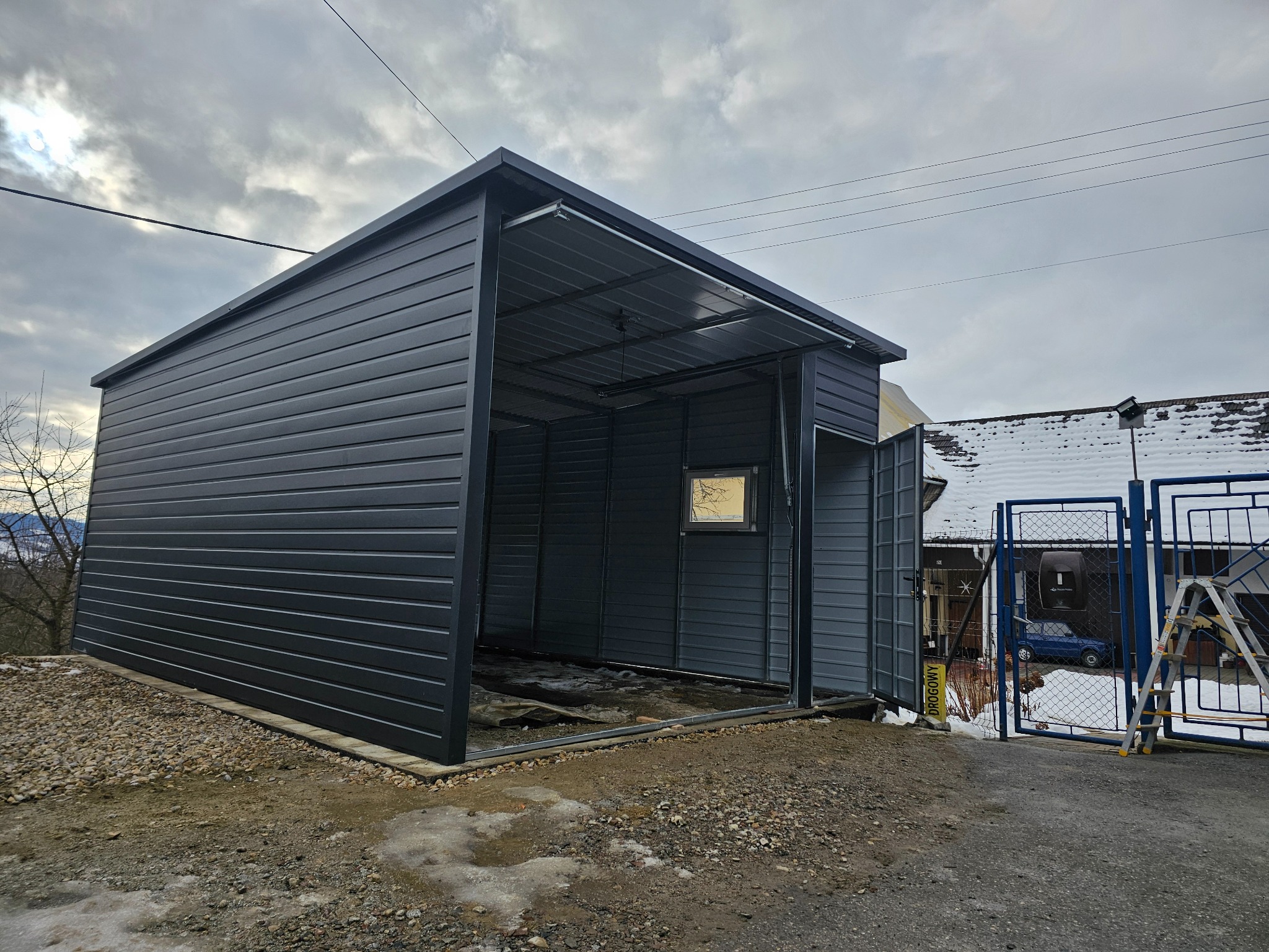 Czarny garaż z blachy trapezowej z podnoszoną bramą, widok z zewnątrz, otoczenie w trakcie przygotowywania, w tle dom z zaspami śniegu na dachu i niebieski samochód za ogrodzeniem.