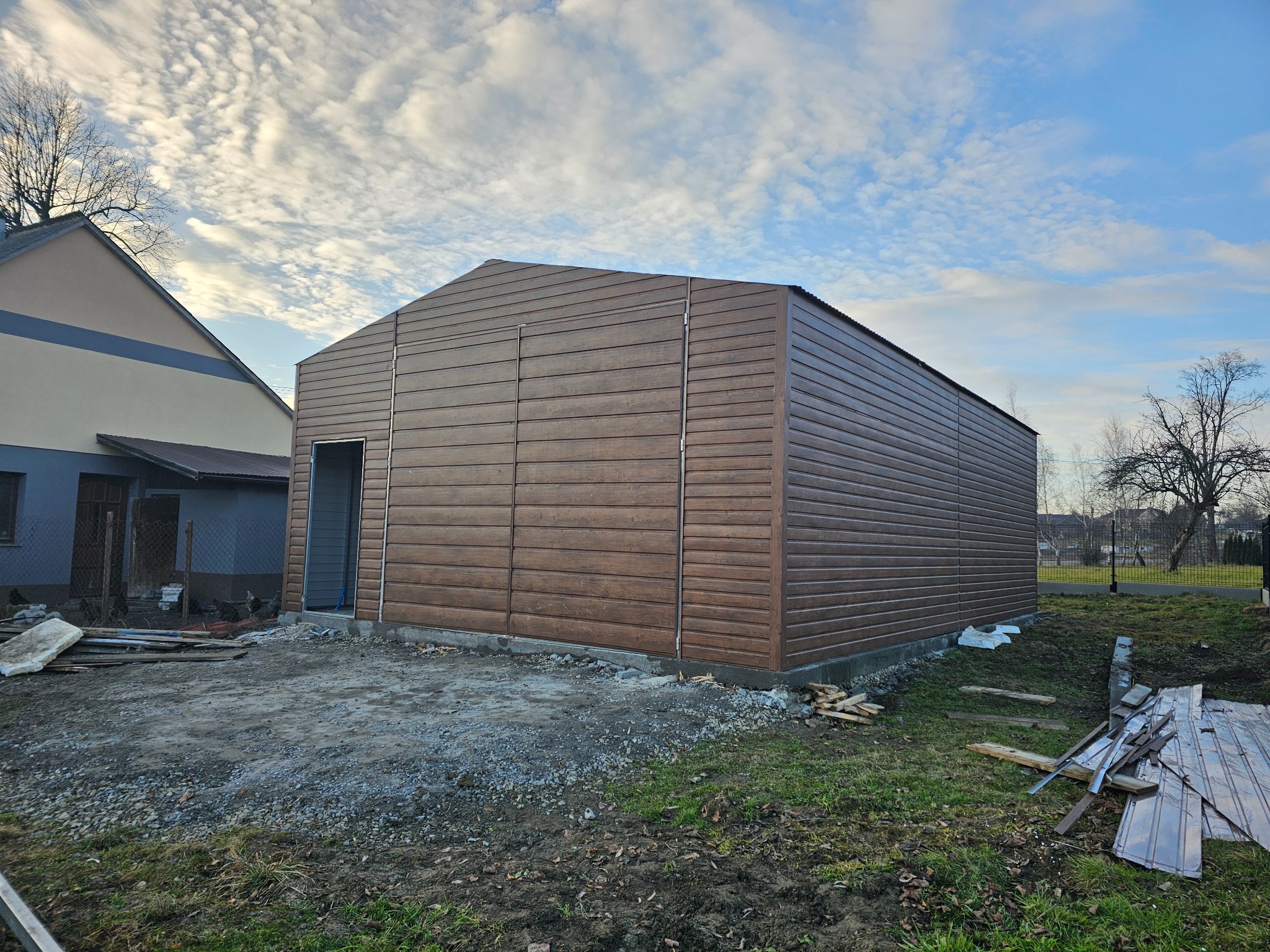 Brązowy garaż z paneli blaszanych z otwartymi drzwiami wejściowymi, widoczne ślady budowy i przygotowane drewniane elementy konstrukcyjne na trawie, w tle budynek mieszkalny i pochmurne niebo.