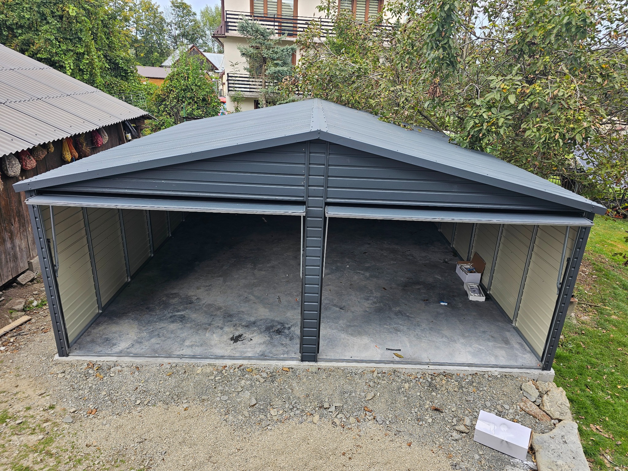 Nowo postawiony, dwustanowiskowy garaż z szarej blachy, z otwartymi wrotami, widok z zewnątrz na żwirowe podłoże i częściowo zarośnięte otoczenie.