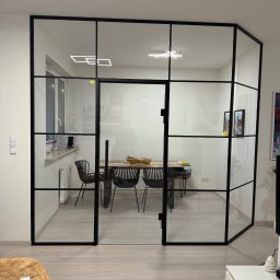 EFEKT SZKŁA Mateusz Wójcik - Nowoczesne drzwi wewnętrzne szklane z czarnymi szprosami oddzielają salon od jadalni. Widoczny stół i krzesła w jadalni, minimalistyczny wystrój.
