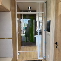 EFEKT SZKŁA Mateusz Wójcik - Nowoczesne drzwi z białym szkieletem i przezroczystym szkłem, z widokiem na zieloną ścianę w tle. Minimalistyczny design w jasnym wnętrzu.